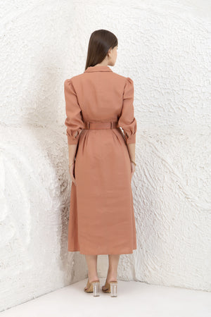 Woman wearing a long brown coat against a textured white wall.
