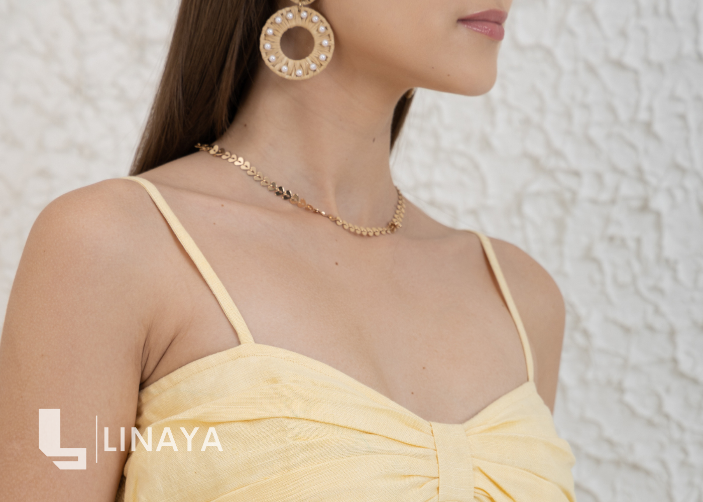 Woman wearing a yellow dress with gold jewelry against a textured white wall, featuring the LINAYA brand.