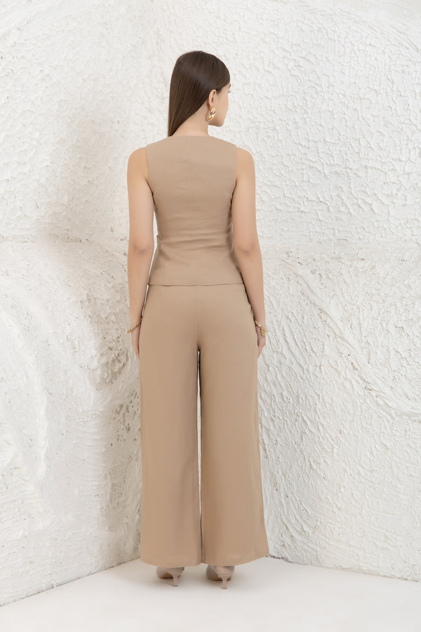 Woman wearing a beige vest and trousers against a textured white wall.
