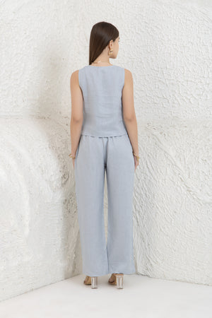 Woman wearing a light blue sleeveless top and wide-leg pants against a textured white wall.