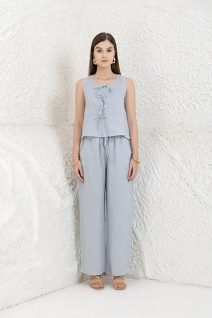 Woman wearing a light gray sleeveless top and wide-leg pants against a white textured wall.