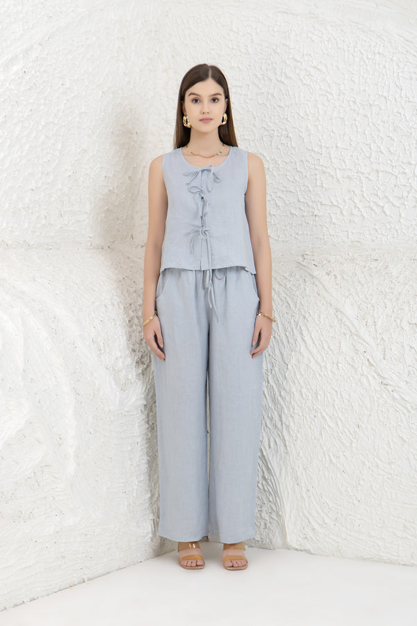 Woman wearing a light gray sleeveless top and wide-leg pants against a white textured wall.