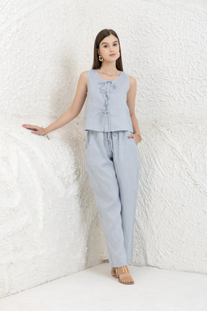 Woman wearing a light blue sleeveless top and pants against a white textured wall.