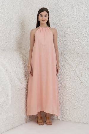 Woman wearing a long pink linen dress women against a white textured wall.