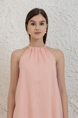 Woman wearing a pink halter neck sleeveless linen dress dress against a textured white wall.